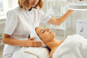 A female cosmetic dermatologist directs a lamp towards a woman's face for a facial treatment.